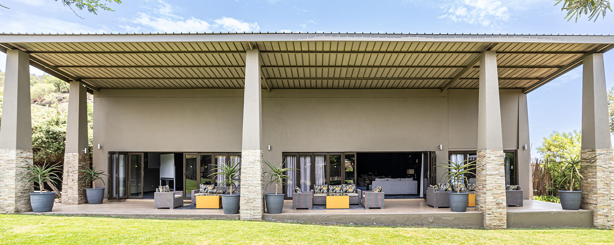 Lodge facade and deep covered veranda with stone-clad pillars, wicker lounge seating, potted aloes and folding glass doors onto a lawn