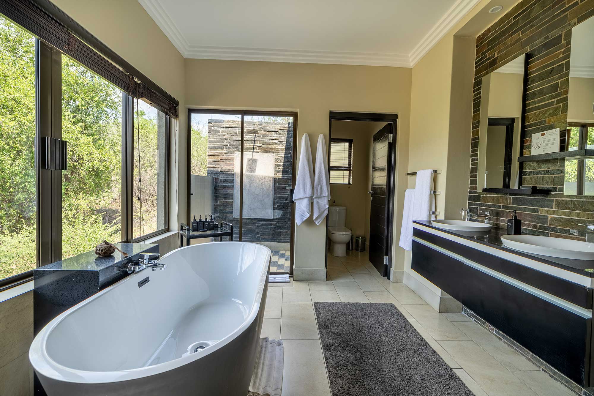 Ensuite bathroom with a freestanding oval soaking tub, a floating double vanity against a stacked-stone wall and windows over the bushveld