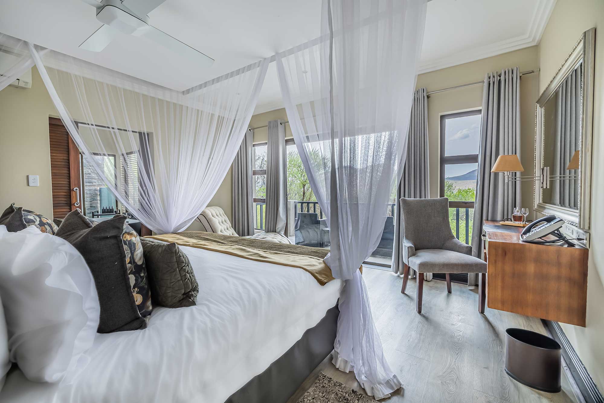 Lodge bedroom with a canopied bed dressed in white linen and netting, an armchair and a wall-mounted wooden desk opening onto a private balcony