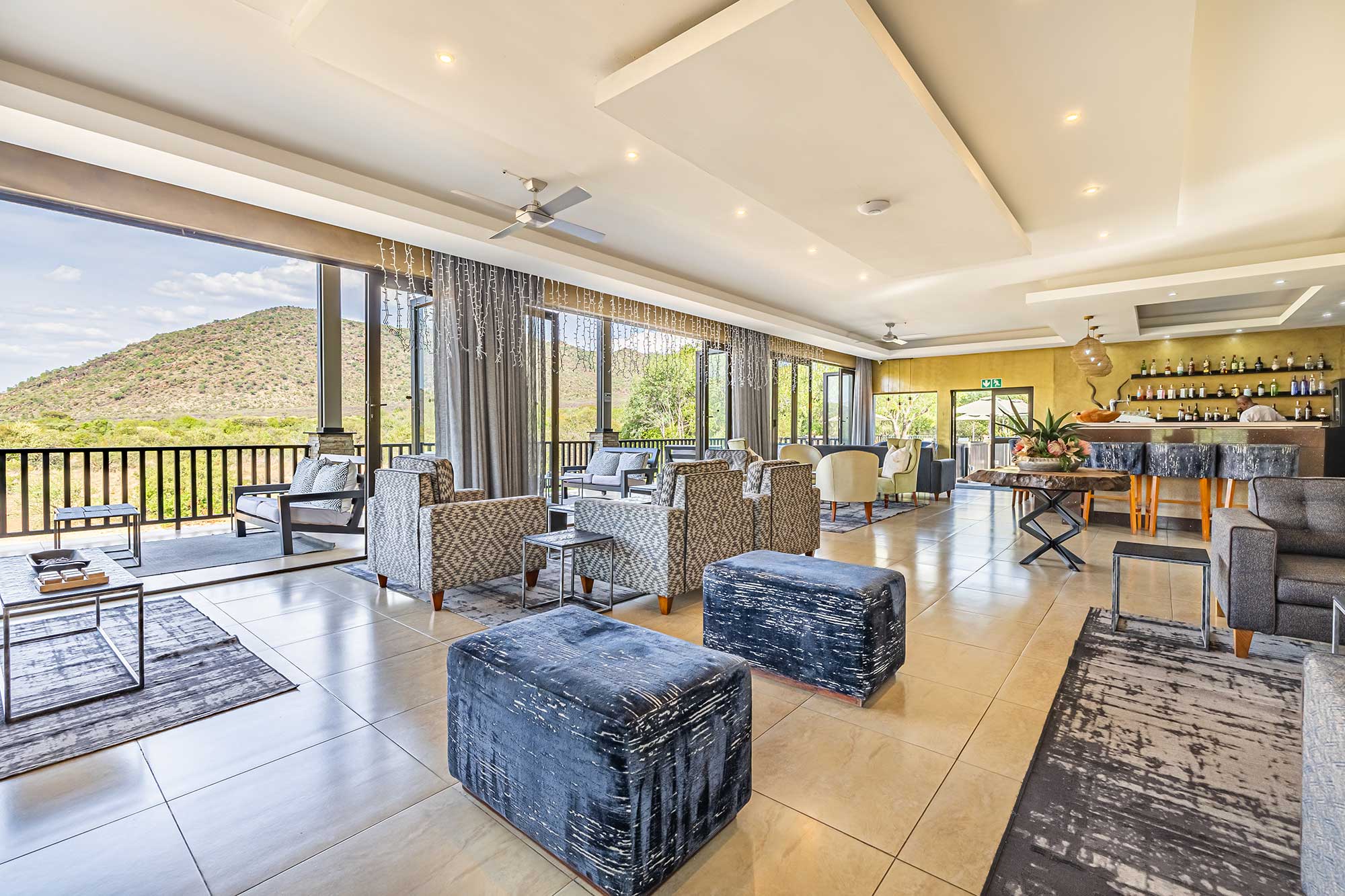 Open-plan lodge lounge and bar with patterned armchairs and blue ottomans, folding glass walls opening to a balcony over the hillside