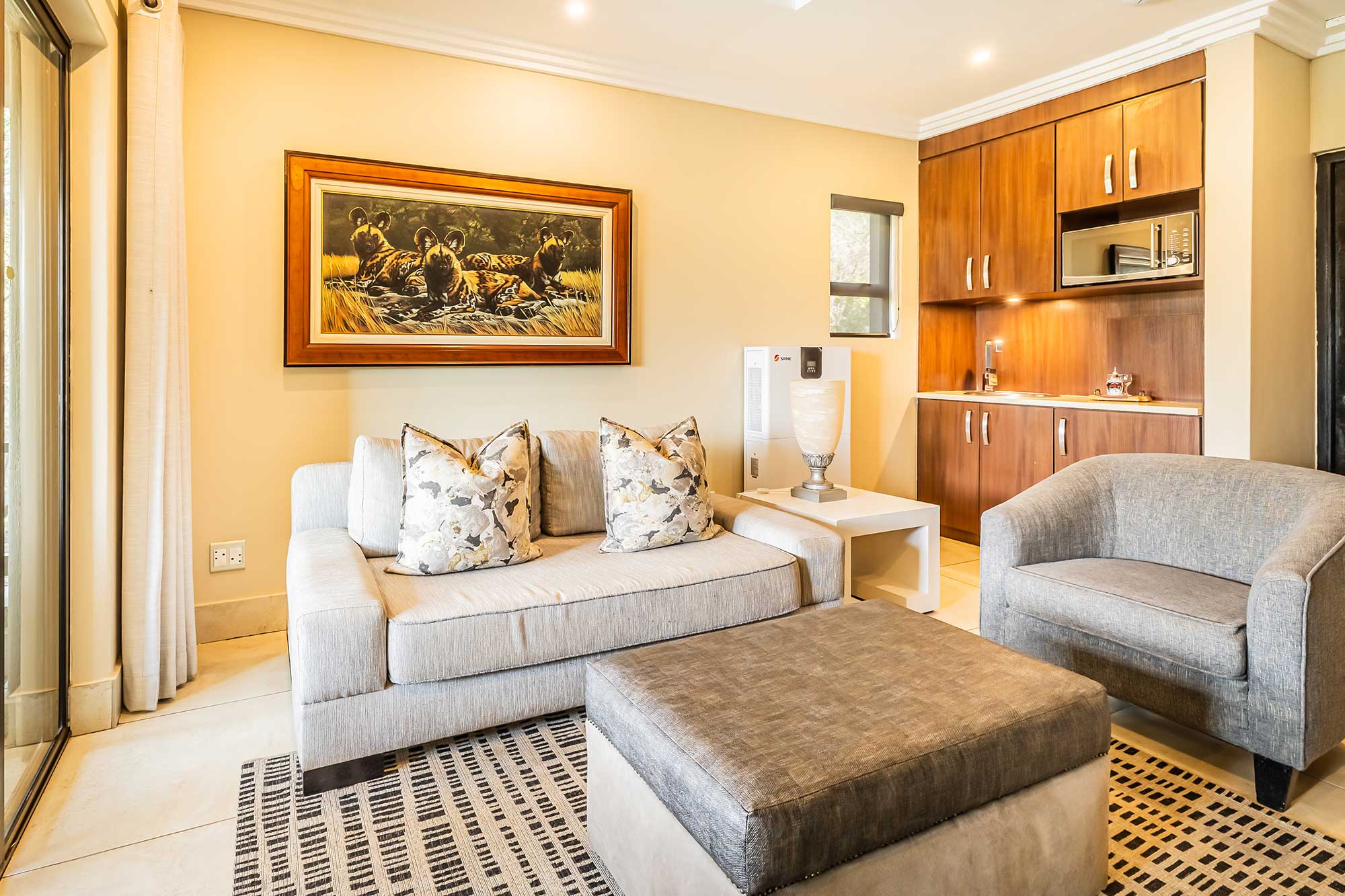 Guest suite lounge with a two-seater sofa, armchair and ottoman, a framed painting of African wild dogs and built-in kitchenette cabinetry