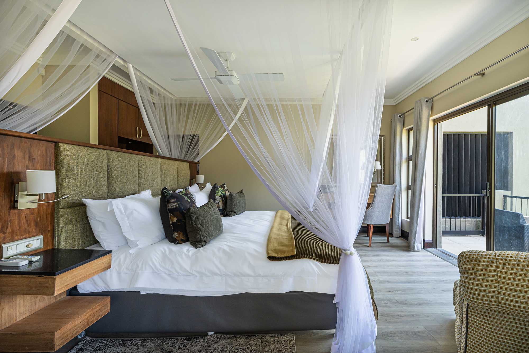 Lodge bedroom with a tall olive-green tufted headboard, suspended white netting and floating bedside shelves opening onto a patio