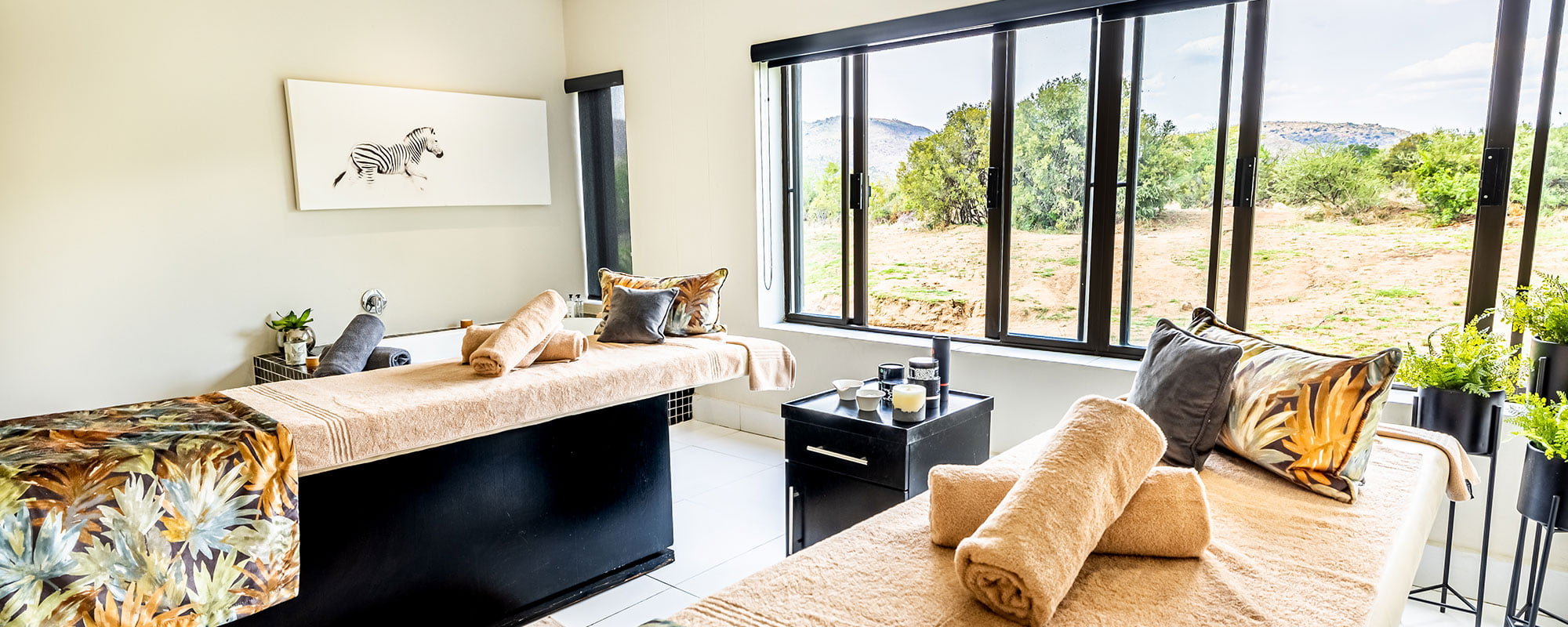 Spa treatment room with two massage beds dressed in towels, panoramic windows onto the landscape and zebra artwork on the wall