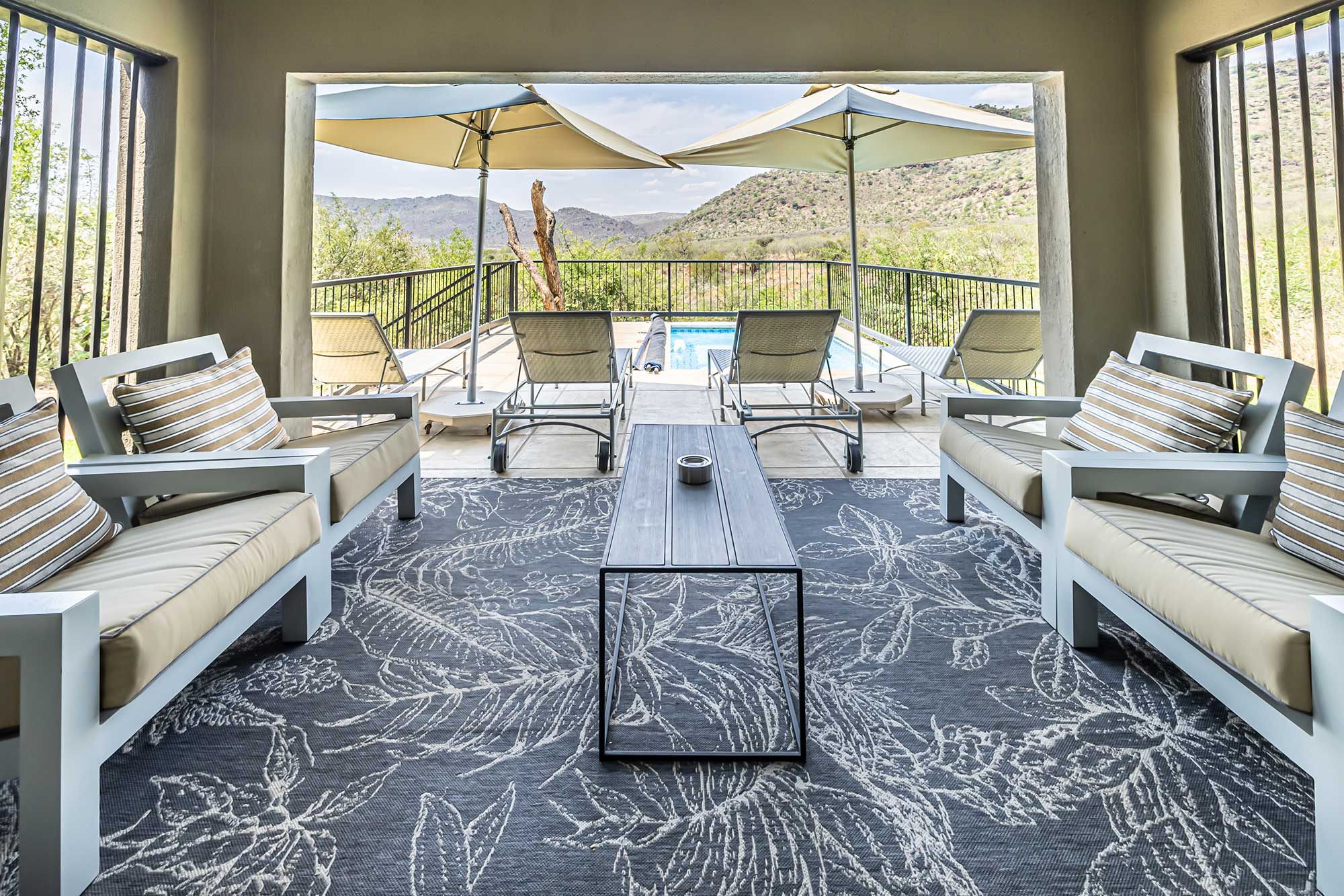 Covered patio with armchairs and a botanical-patterned rug framing a view of the pool deck, sun loungers and distant hills