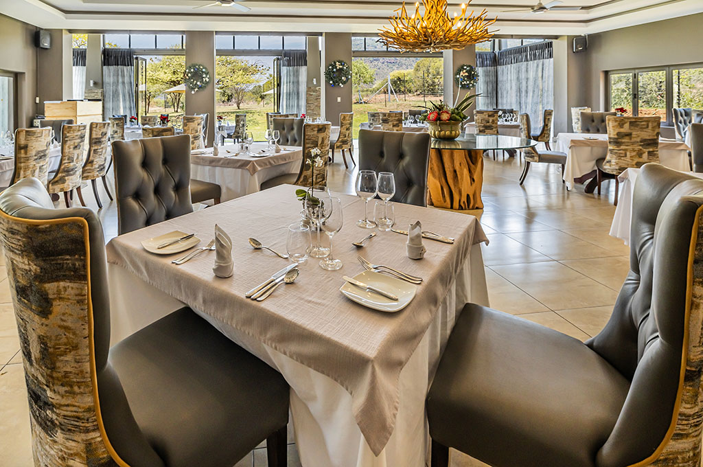 Lodge dining room laid with linens, glassware and cutlery, tufted high-back chairs, a sculptural ceiling light and windows over the landscape